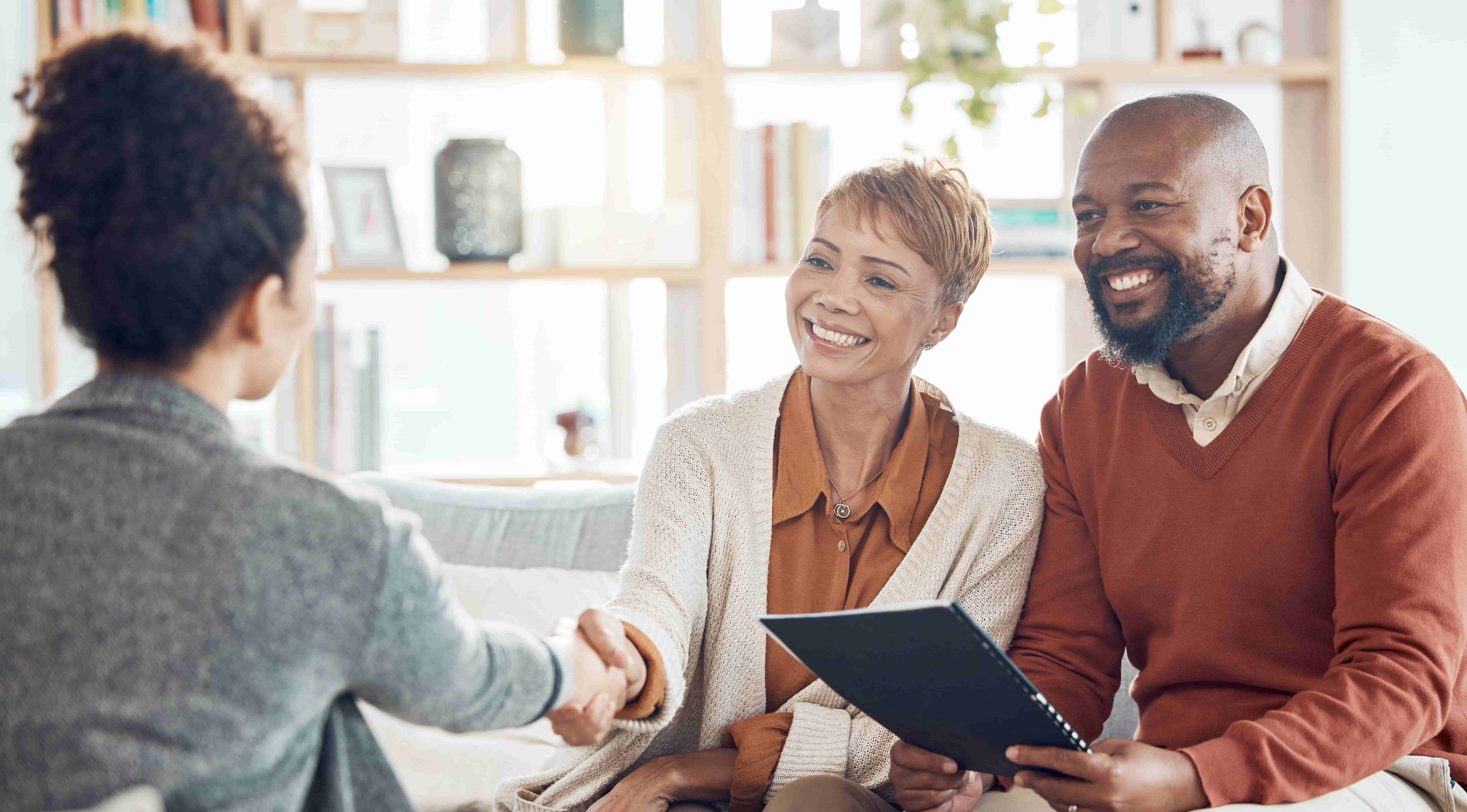 Smiling couple meets with a professional advisor at home. The woman shakes hands with the advisor while the man holds a folder, all three engaged in a warm and positive conversation.