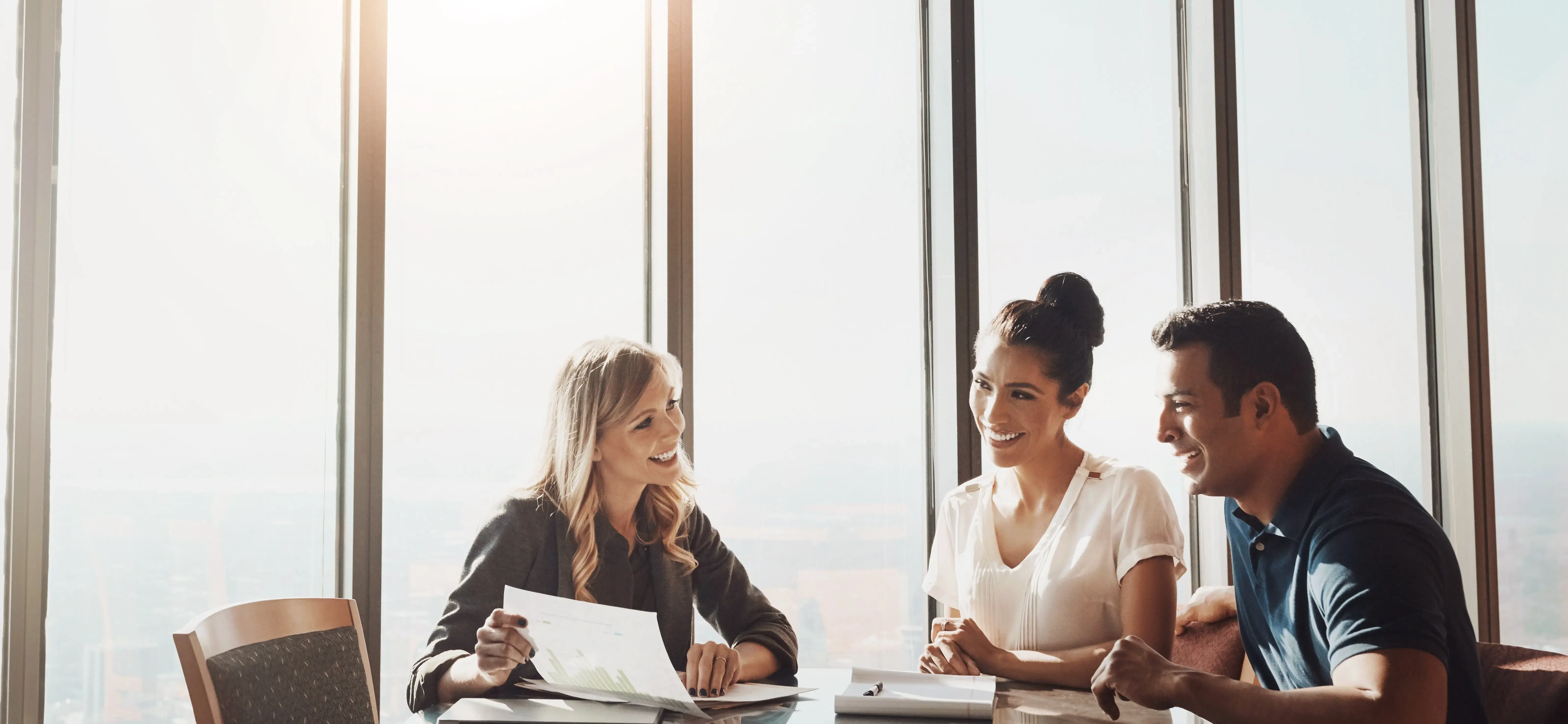 Financial advisor meeting with a smiling couple in a bright office, discussing documents across a glass table.