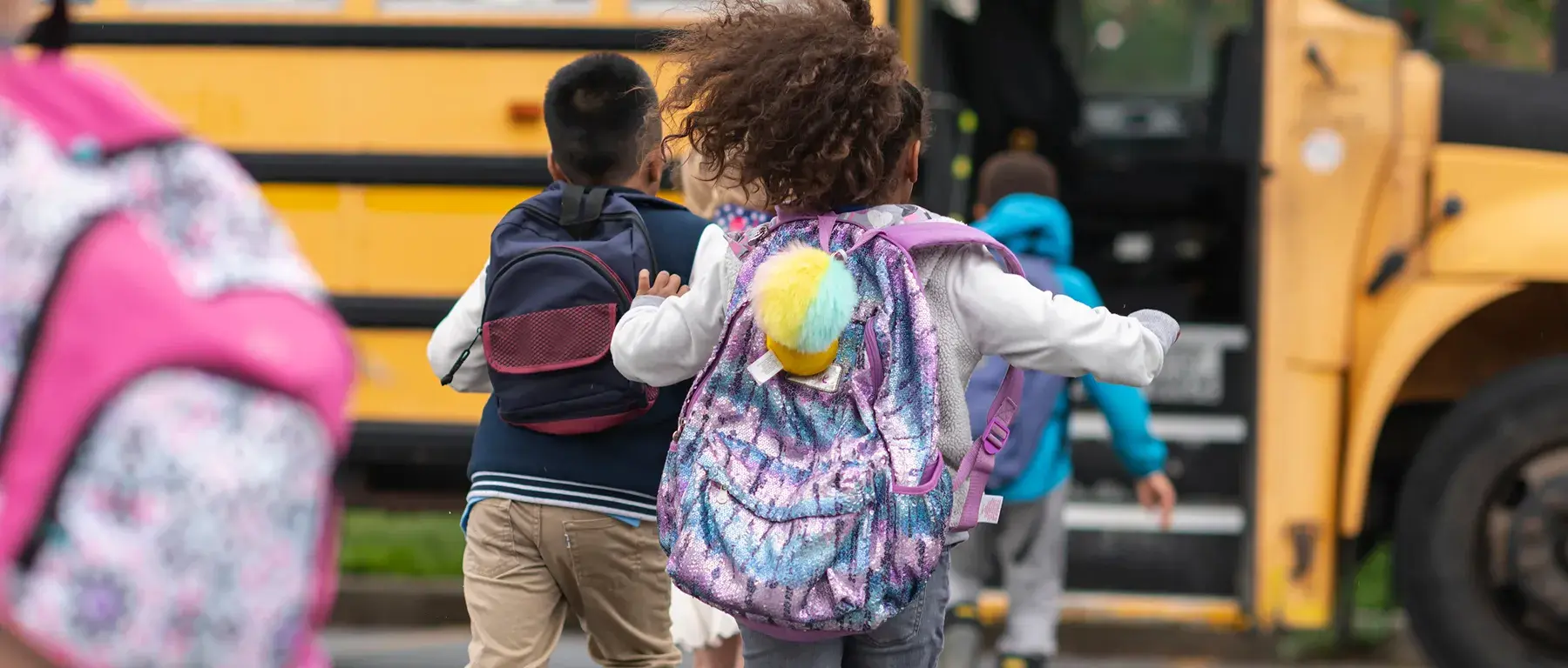 Children with backpacks walking toward a yellow school bus, preparing to board.
