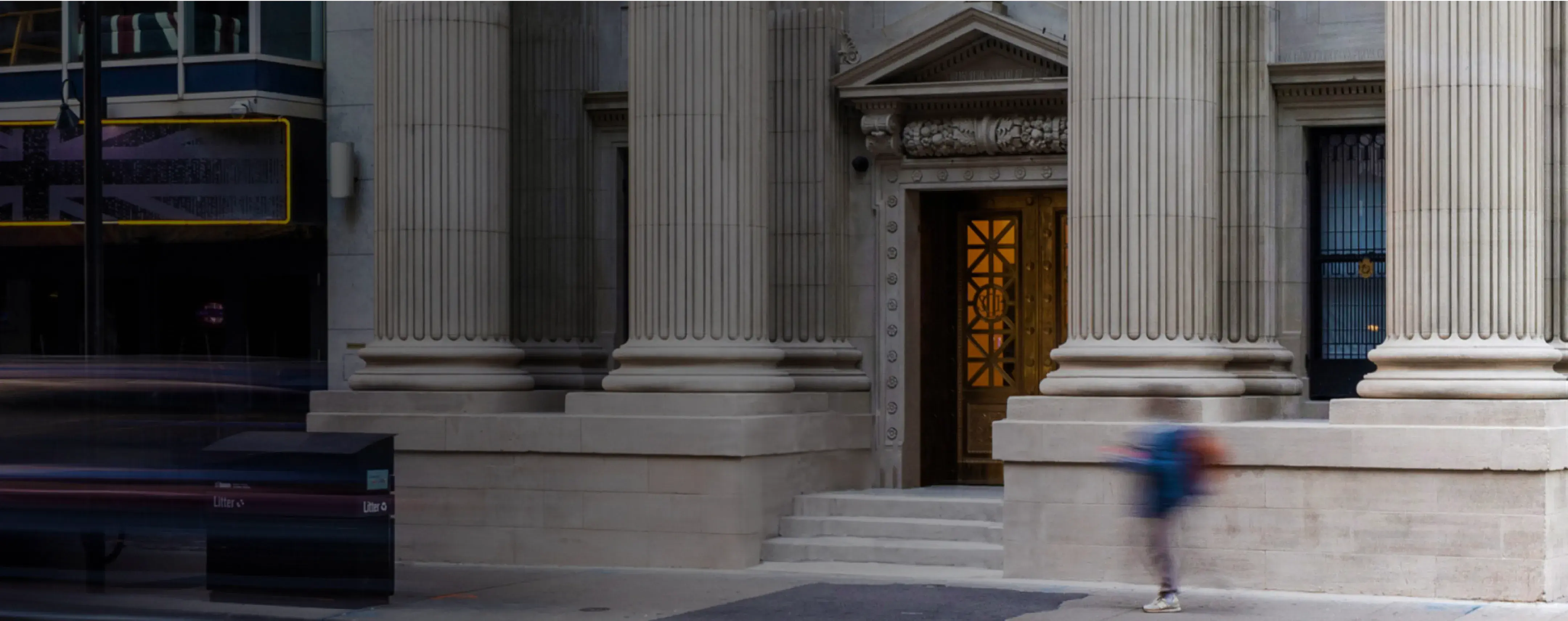 Entrance of a neoclassical building with tall stone columns and ornate golden doors, with a blurred pedestrian walking past in the foreground.