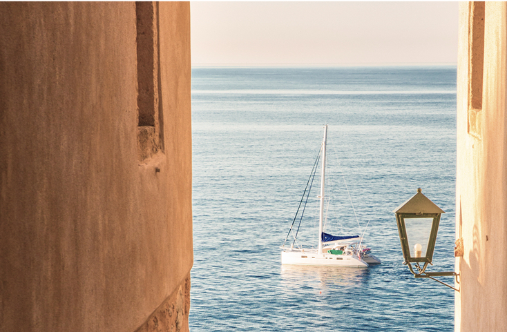 Small sailboat on calm blue sea, viewed through narrow stone alley walls.