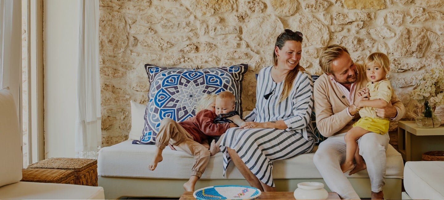 Smiling couple sitting on a sofa with their young child, in a cozy, sunlit room.