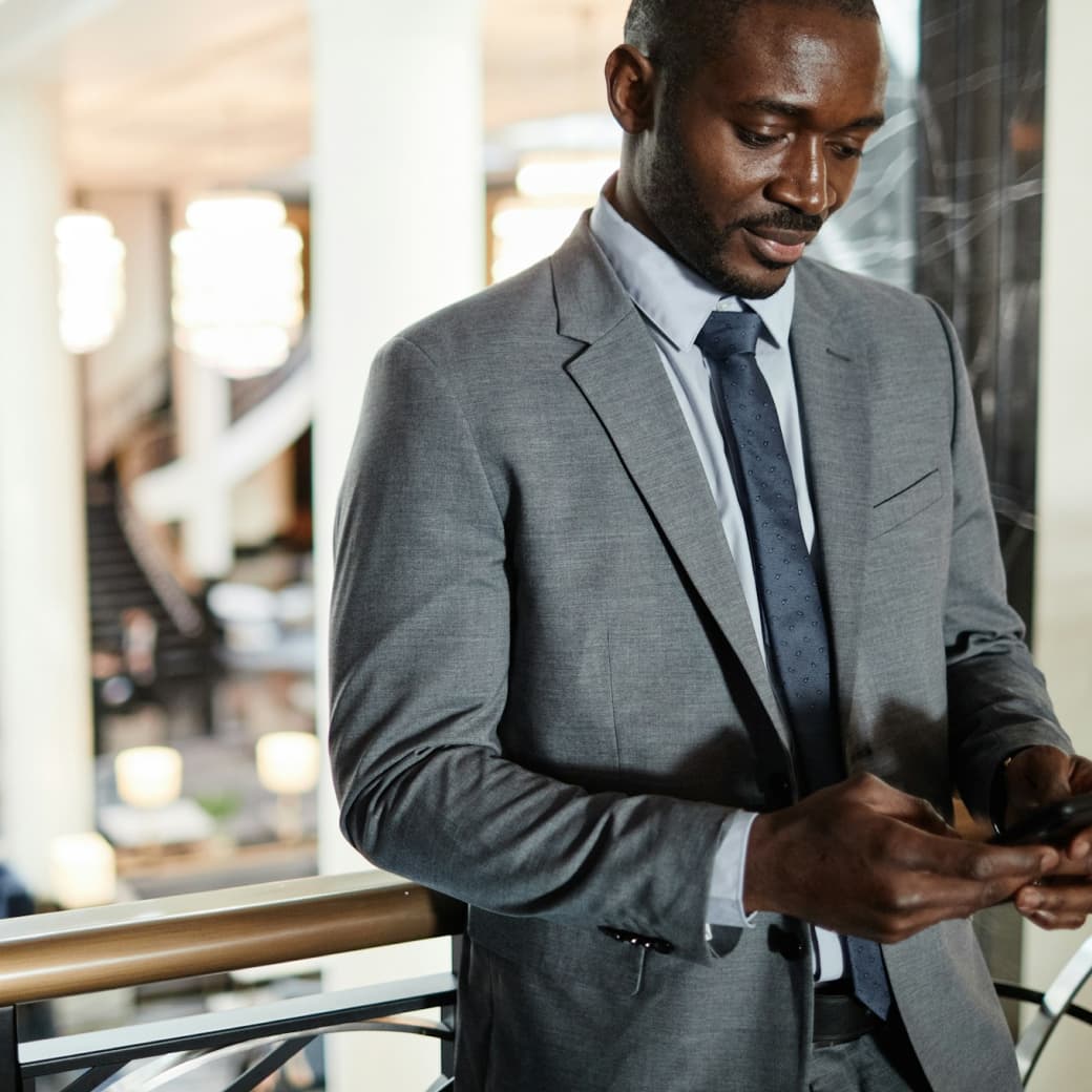 A man in a grey suit and blue tie standing indoors, leaning against a railing while looking down at his phone with a slight smile.