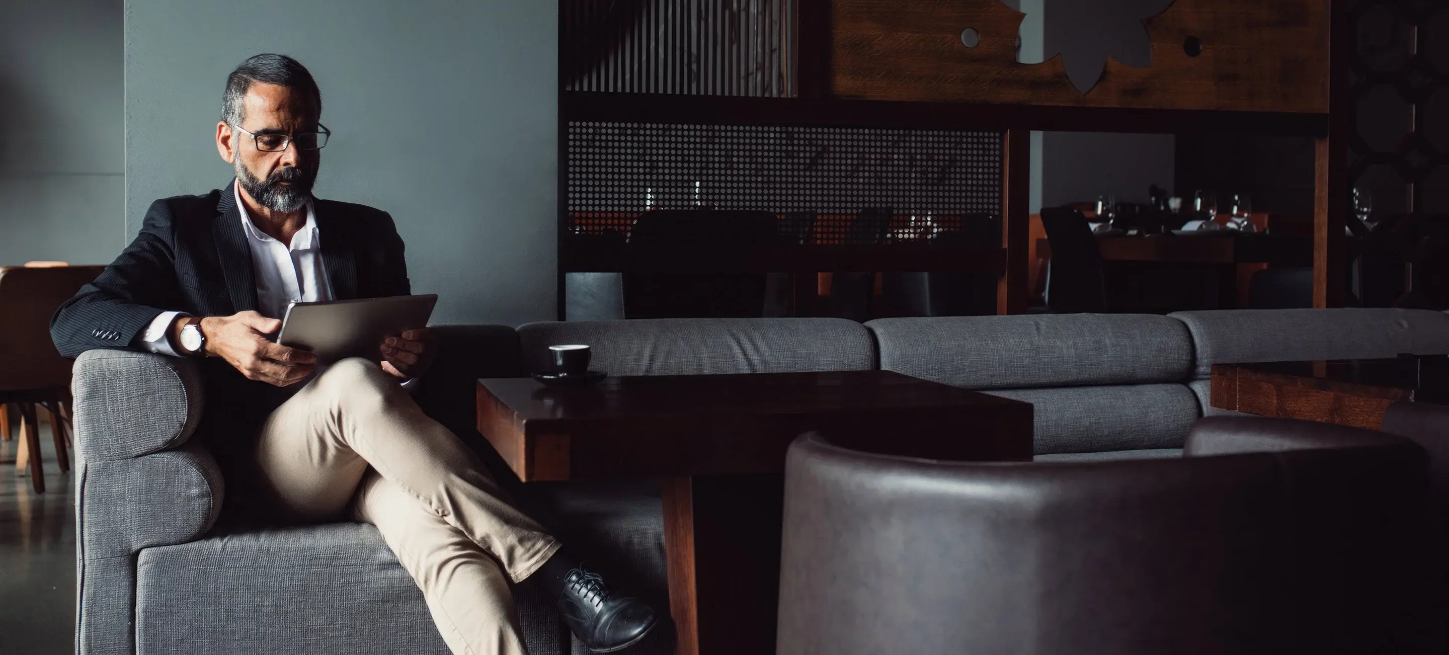 Man sitting on a sofa in a lounge, wearing a dark jacket and light pants, reading a document.