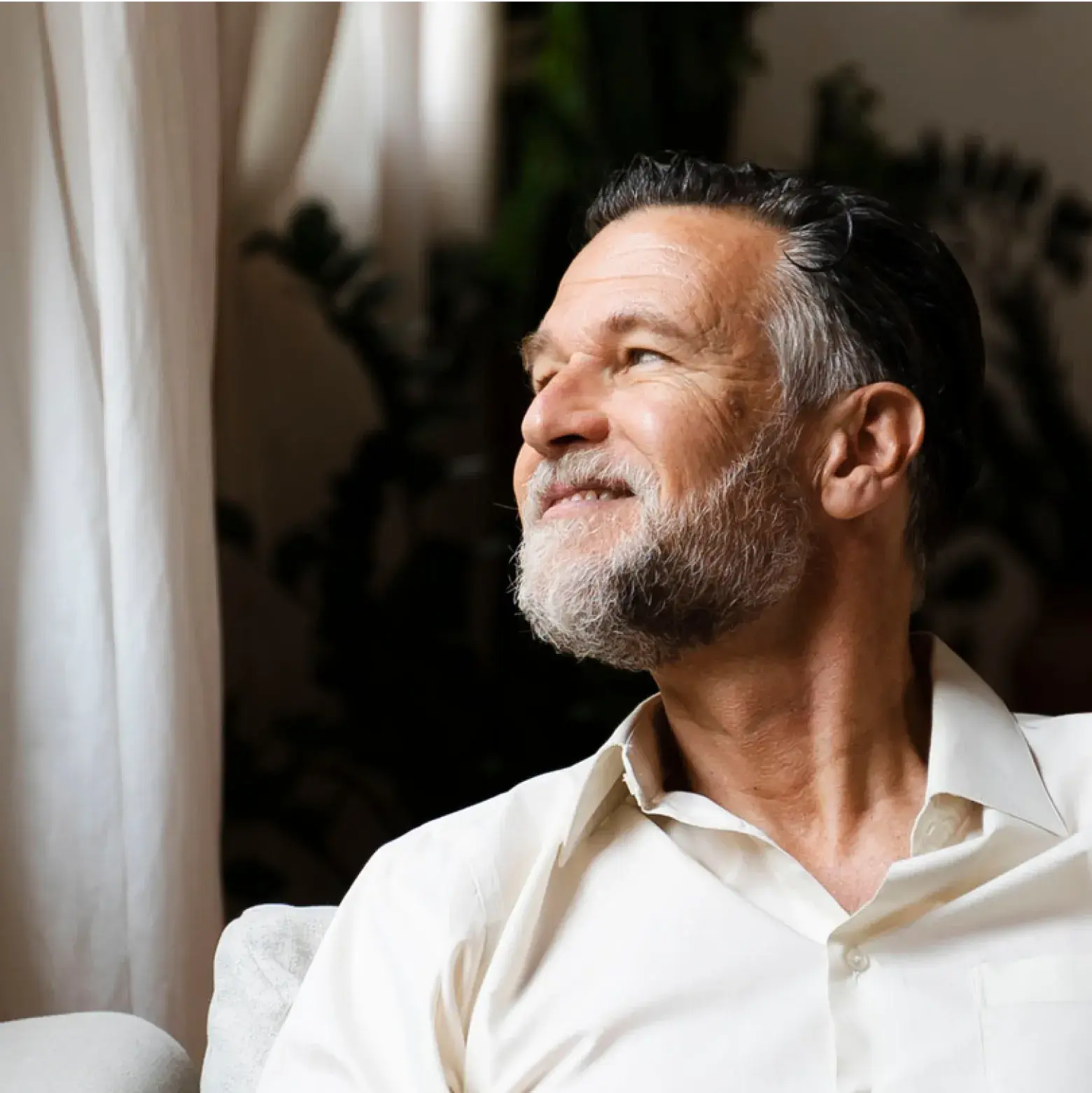 Smiling older man with gray hair and beard, wearing a light shirt, sitting indoors and looking toward the window with soft natural light on his face.