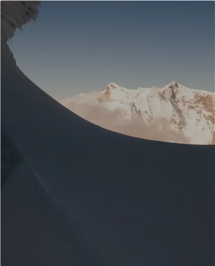 Snow-covered mountain ridge in shadow, framing a distant sunlit peak against a clear blue sky.