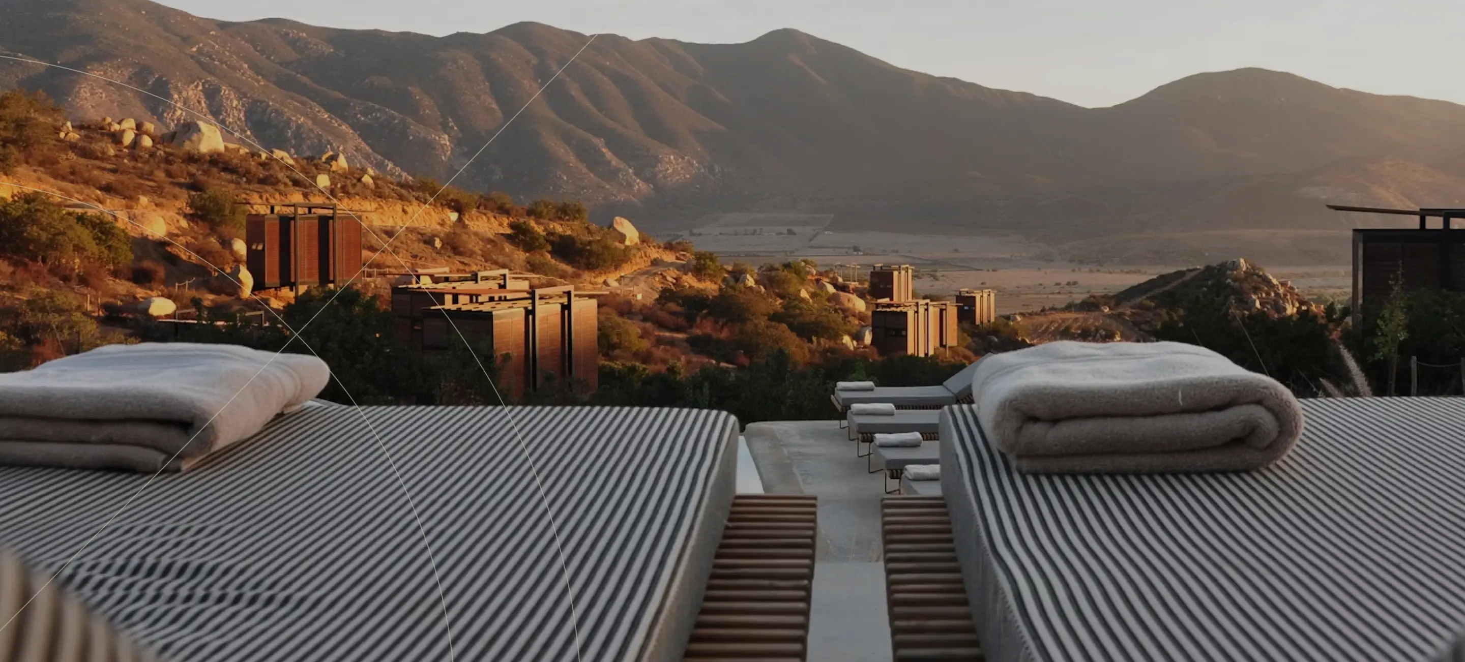 Outdoor lounge chairs with folded blankets overlooking a valley of modern wooden cabins, set against sunlit mountains in the distance.