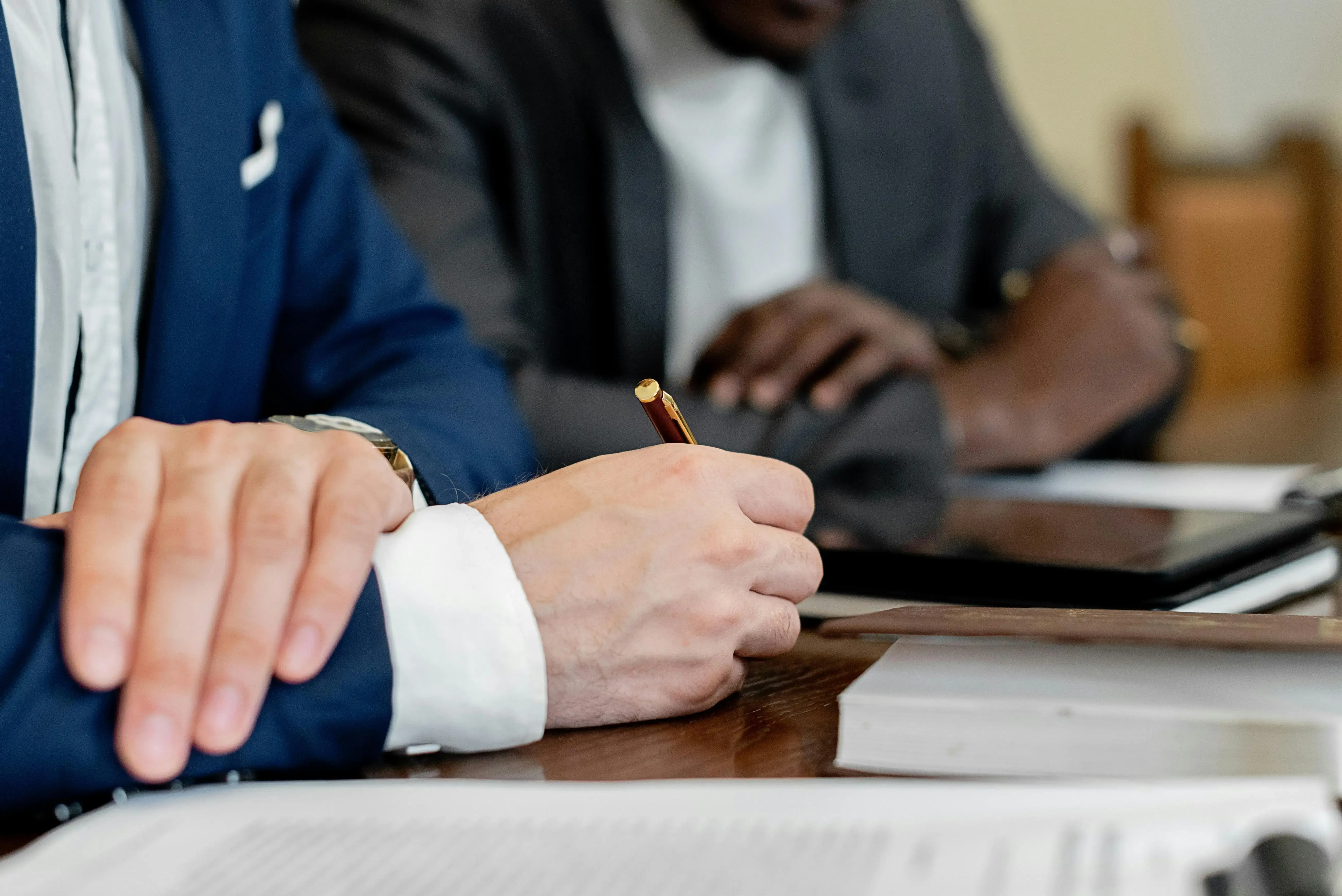 Two professionals in formal business attire sit at a conference table. One person in a blue suit is writing with a pen on paper, while another in a dark suit listens beside a tablet. Documents and books are spread across the table, suggesting a meeting or financial discussion.